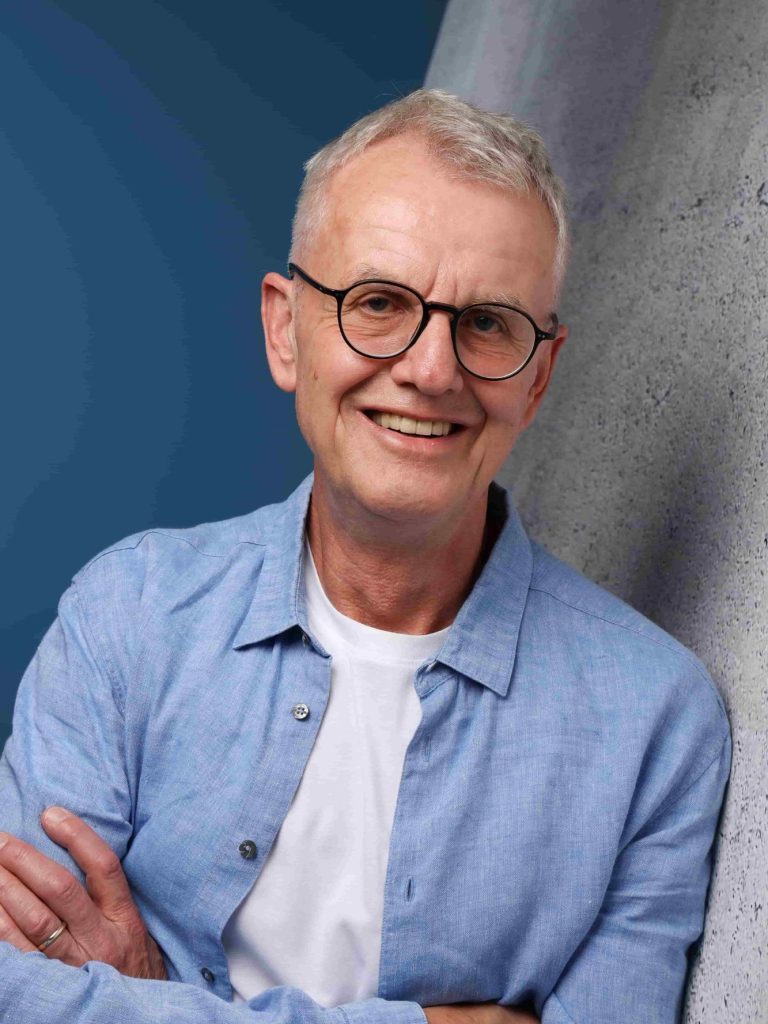 Ein lächelnder Mann mit Brille, kurzen grauen Haaren und in lässiger Kleidung.