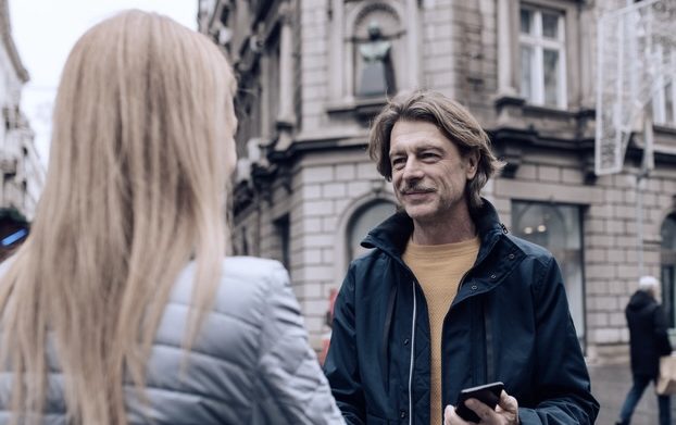 Mann mittleren Alters spricht auf der Straße mit Frau, lange blonde Haare, nur ihre Rückseite ist zu sehen.