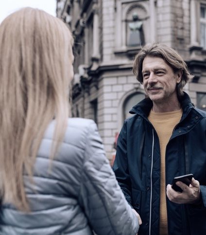 Mann mittleren Alters spricht auf der Straße mit Frau, lange blonde Haare, nur ihre Rückseite ist zu sehen.
