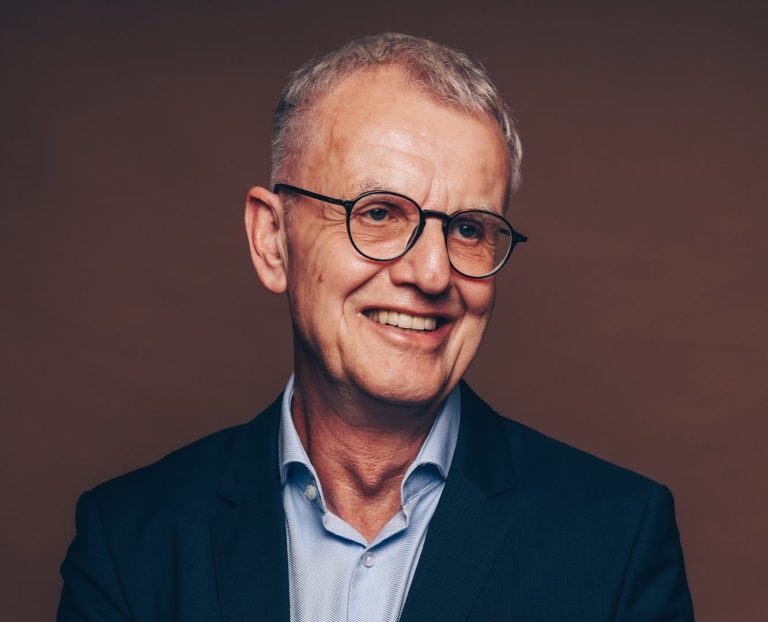 Mann mit Brille und silbernem Haar, lächelnd in Anzug mit gekreuzten Armen.