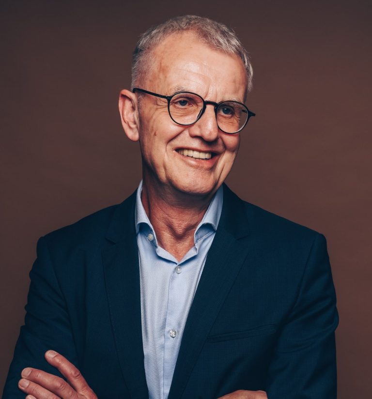 Hochschuldozent und Medientrainer Mann mit Brille und silbernem Haar, lächelnd in Anzug mit gekreuzten Armen.